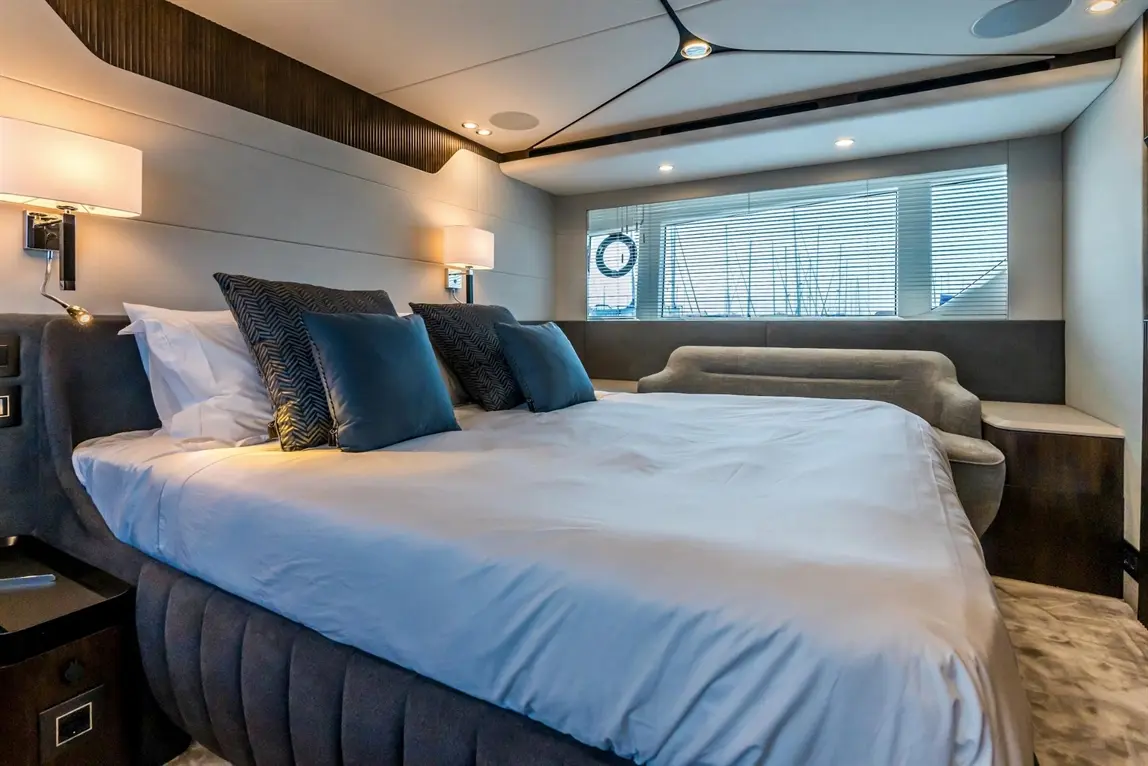 A modern bedroom featuring a large bed with blue cushions, stylish lighting, and a window with blinds overlooking a marina.